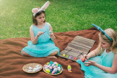 Cute little girls wearing bunny ears on Easter day sitting on blanket on grass in garden and...