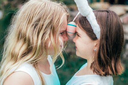 Cute little sisters wearing bunny ears on Easter day d playing with painted egg holding it between...