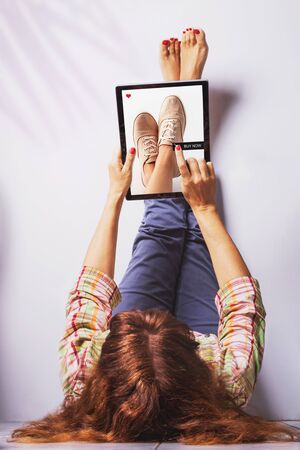 Woman lays with legs up on the wall and pushes buy now button on a laptop buying shoes. Online shopping concept.Self isolation.の写真素材