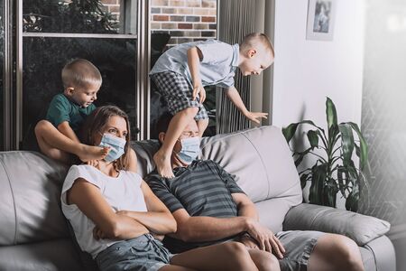 Mom and dad are wearing facial surgical masks. Two little sons are jumping over parents sitting on the couch trying to watch TV. All family stays together at home during quarantine lockdown.の写真素材