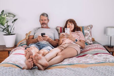 Middle-aged couple using their mobile phones laying on bed with their legs together. Internet social network addiction concept.の写真素材