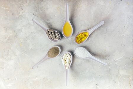 Capsules fish oil omega-3, white and gray pools, oil and powder in spoons in a circle on the white rustic background. Health and beauty concept.の写真素材