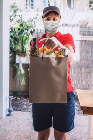 Delivery woman in protective face mask and gloves delivers paper bag with food products. Health protection, safety and pandemic concept.の写真素材