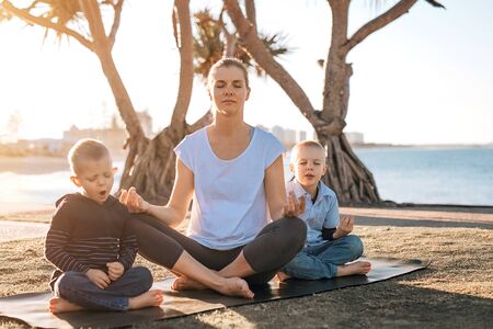 Woman meditating outdoors in park with two cute little sons. The ocean on background. Selective focus.の写真素材