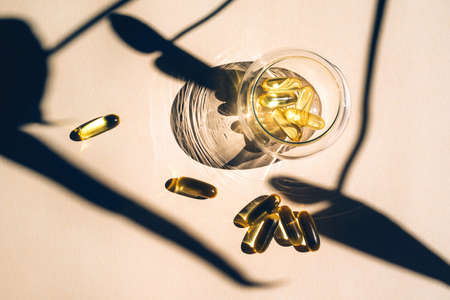 Capsules fish oil omega-3 in glass cup with a shadow of monstera leaves in the sunlight on the beige background. Health and beauty concept.の写真素材