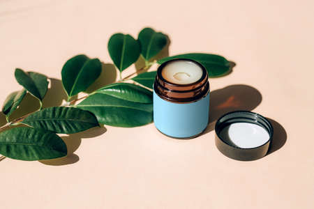 Jar of essential oil cream with natural plant leaves on a beige background.