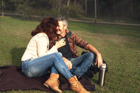 Happy middle-aged couple in love sitting on a blanket in the grass in park, hug, look at each other,...