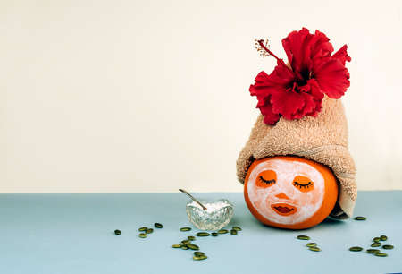 Pumpkin with painted face and facial mask, towel with flower, pumpkin sseeds and powder in glass bowl on pastel blue background. Spa concept, Helloween, Copy space.の写真素材