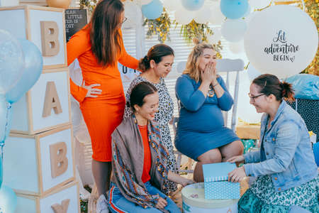 Female friends open gift boxes with a pregnant woman at a baby shower. Party decorations in white...