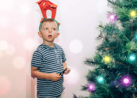 Cute little kid with deer antlers on the head playing with fairy lights on Christmas tree.の写真素材
