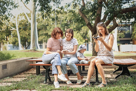 Three women friends with baby strollers reading text message in parkの写真素材