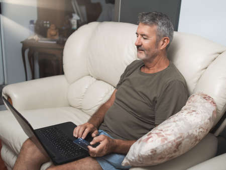 Smiling middle aged man sitting on the couch using the laptop to buy online at homeの写真素材