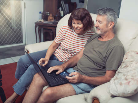 Smiling middle aged couple sitting on their couch using the laptop to buy online at homeの写真素材