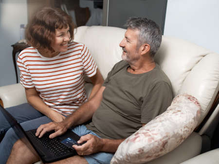 Smiling middle aged couple sitting on their couch using the laptop to buy online at homeの写真素材