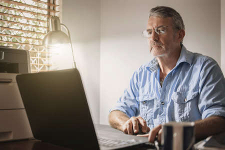 A senior man sitting at a desk in front of a laptop computerの写真素材