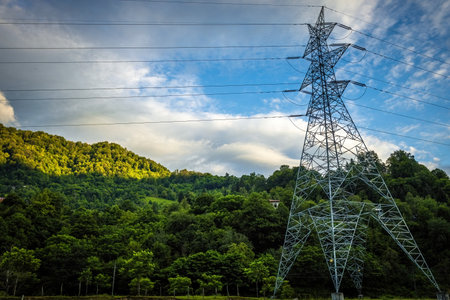 High-voltage electricity transmission tower standing in a lush green forested landscape with hills and dramatic clouds in the backgroundの写真素材