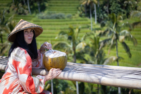 Young beautiful woman wearing asian hat and drinking coconut sitting on the rice fields in Baliのeditorial素材