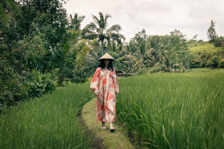 Woman walking along the green rice fields wearing long red and white dress and Asian conical hat in...
