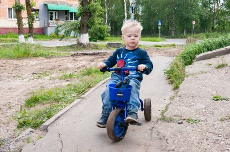 Russia, Novodvinsk - July 2019. Blond-haired boy in blue rides a small tricycle on a broken city pathのeditorial素材