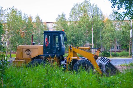 Road repair work - an orange excavator stands on the road behind a green wall of grass against a...