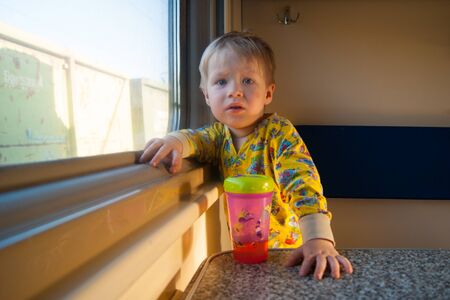 A bright cute three-year-old boy in yellow pajamas rides on a train by the window, behind which a...
