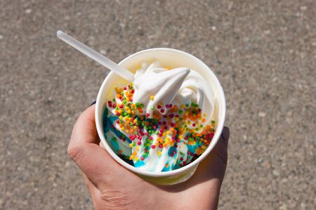 Female hand holds soft tender ice cream with colored topping and syrup in a paper cup