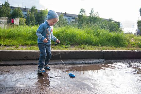 A three-year-old white boy in a blue hat, jeans and sandals plays on a cool summer day with an outdated remote control car. The kid stands sideways to the viewer on a background of green grass.の写真素材