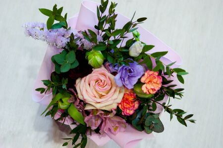 A delicate bouquet of different flowers in a light pink wrapper stands on a light background in a...