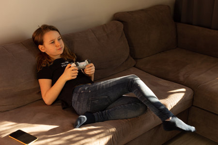 A girl in jeans and a T-shirt is playing video games at home on the couch, there is an inscription...