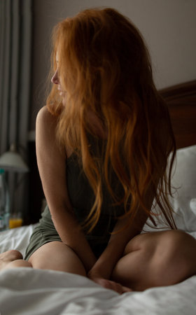 Sexy woman with red hair sitting on the bed in the morning light from the windowの写真素材