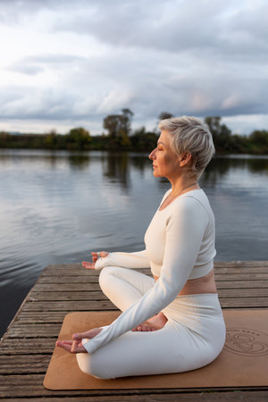 Mature woman in a white suit in a lotus position on the river bankの写真素材