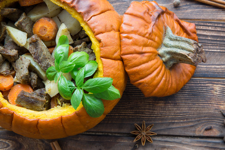 Baked meal in pumpkin on rustic brown wooden background. Stewed potatoes with carrot and roasted meat (veal and pork), decorated with basil leaves. Autumn mood, thanksgiving fall background.の写真素材