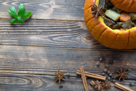 Baked meal in pumpkin on rustic brown wooden background. Stewed potatoes with carrot and roasted meat (veal and pork), decorated with basil leaves. Autumn mood, thanksgiving fall background. Copy space.の写真素材