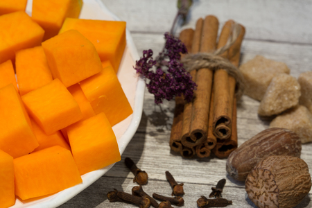 Chopped pumpkin on white plate with cinnamon sticks, anise star, nutmeg and cloves on grey wooden table background. Ingredients for baking tasty pie or cupcakes.の写真素材