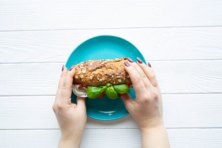 Woman hands holding delicious healthy sandwich with bacon, vegetables and whole wheat bread on white wooden table, top view.の写真素材
