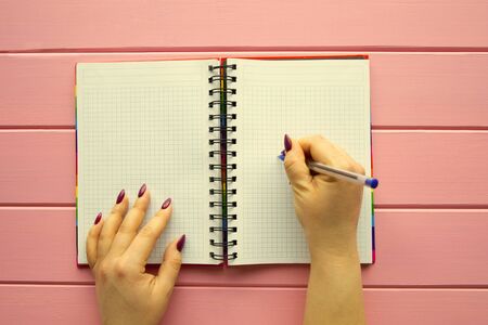 Female hands is writing something in the blank notebook on pink colorful background, top view. Plan...