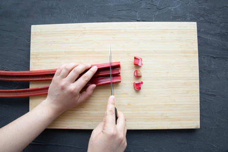Fresh rhubarb on wooden cutting board on black background. Female holds knife and chop, top view.の写真素材