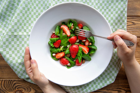 Delicious fruit summer salad, female hands hold plate and fork, rustic wooden background, top view.の写真素材