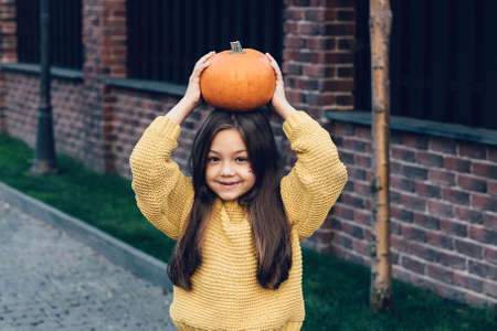 Funny child girl in orange pullover for Halloween with pumpkin and on a dark brick background. Happy laughing child girl in orange pullover and black jeans to halloween.の写真素材