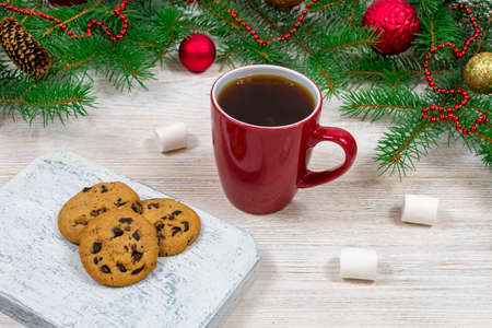 Red cup with tea, coffee with cookies and marshmallows on a white wooden table against the background of a New Year tree with Christmas decorations. Coziness and New Year concept.の写真素材