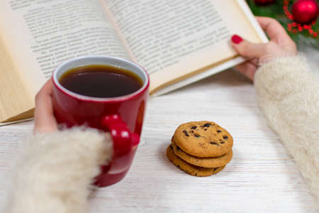 Female hands with a cup of tea and cookies on a wooden table with a Christmas tree and decorations hold a book. Blurred background. Partial focus. Christmas and New Year concept.の写真素材