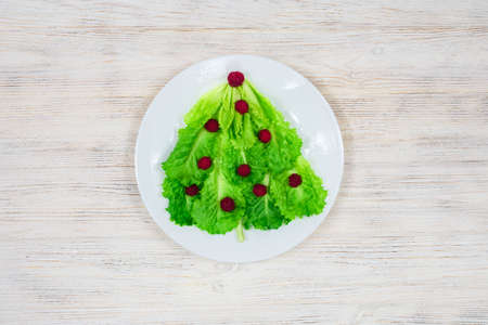 The Christmas tree is lined with green lettuce leaves, decorated with red raspberries on a white plate. Food for the New Year. Table decoration.の写真素材