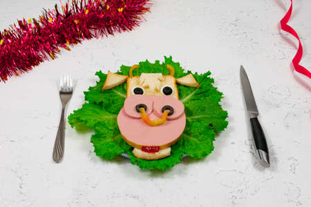 An edible symbol of 2021. Creative children's sandwich in the form of a bull, a cow on a plate. Cute sandwich in the shape of a cow with sausage, olives, cheese, on lettuce leaves. Table, tinsel. New Year's menu.の写真素材