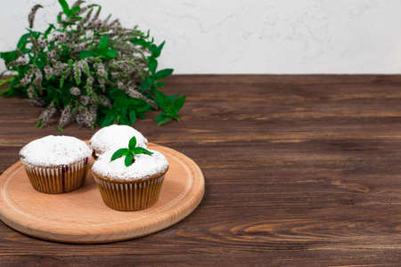 Blackcurrant cupcakes with a white cup of tea on a background of flowers and mint leaves, on a round board on a wooden background.の写真素材