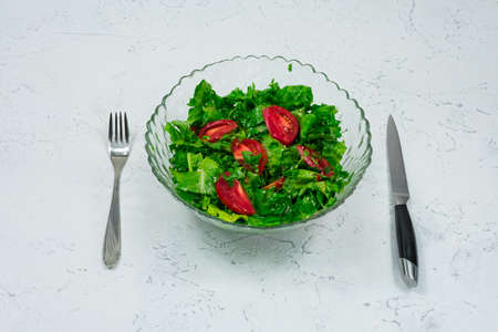 Green vegan salad made from a mixture of green leaves and tomatoes in a salad bowl with a fork and knife on a white stone table.の写真素材