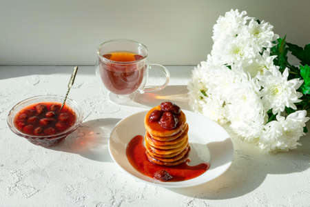 Fritters in a white plate with strawberry jam, a cup of tea and a bouquet of white chrysanthemums on a white table.の写真素材