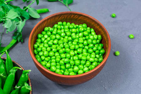 Close-up. Selective focus. Defocus. Bowls of fresh young green peas with stitches and peeled leaves on the background of shoots, sprigs of young green peas on a gray table.の写真素材