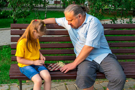 Little girl plays chess with dad in the park on a bench in the fresh air. Chess day. Educational, educational outdoor games. Child development.の写真素材