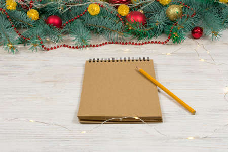 Blank notebook with a white pen on a white table with gold sparkles, stars on the background of a Christmas tree decorated with toys. Festive concept. Place for an inscription.の写真素材