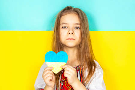 A girl in a national Ukrainian vyshyvanka holds a heart painted in yellow and blue tones as a sign of love for Ukraine, close-up against the background of the Ukrainian flag. Place for text.の写真素材
